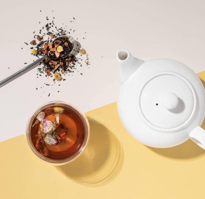 Creative flat lay of a glass cup of herbal tea with rose flowers on a yellow and white background with teapot and hard shadow. Food and drink concept. Creative flat lay of a glass cup of herbal tea with rose flowers on a yellow and white background with teapot and hard shadow. Food and drink concept. Top view and copy space.