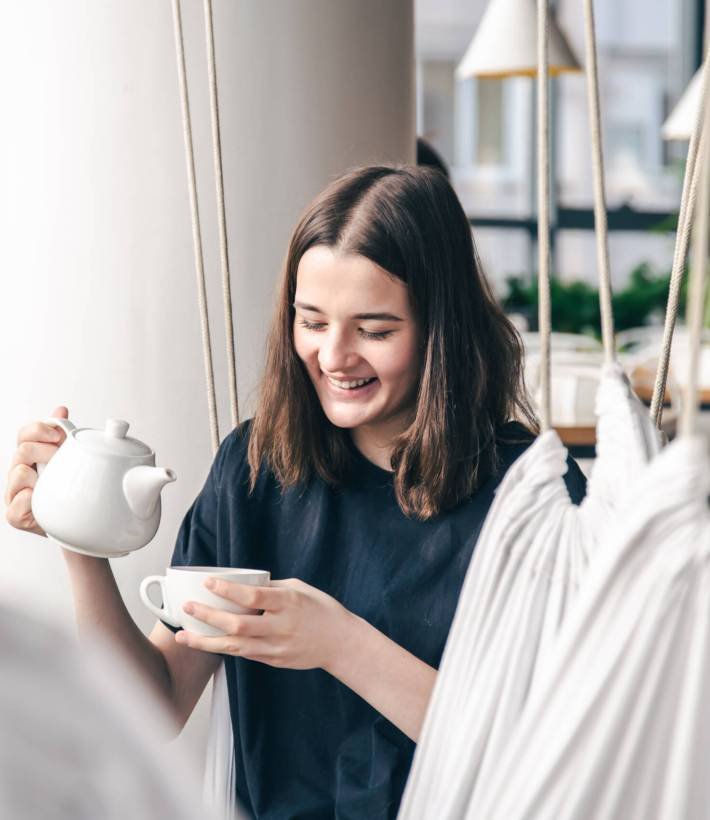 Portrait of a young woman with a cup of tea in a cafe. A happy young woman sits on a hanging hammock in a cafe with a cup of tea and teapot in her hands.